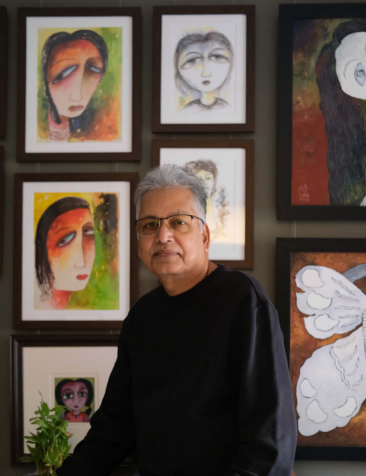 Ratnakar Ojha with gray hair and glasses, wearing a black long-sleeve shirt, stands in front of a wall decorated with multiple of his framed colorful paintings of faces and abstract designs.