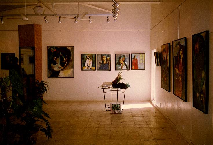 An art gallery interior exhibit of Ratnakar Ojha with multiple framed paintings hanging on beige walls, a central table with a sculpture, and a tiled floor under track lighting.