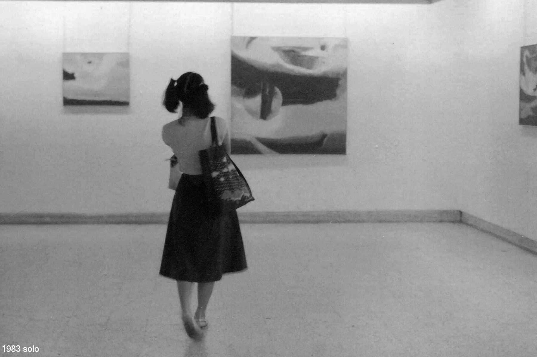 Black and white photograph of a woman at a Ratnakar Ojha exhibit seen from behind looking at a large abstract painting on the wall. She is wearing a sleeveless top and a dark skirt, with a shoulder bag.