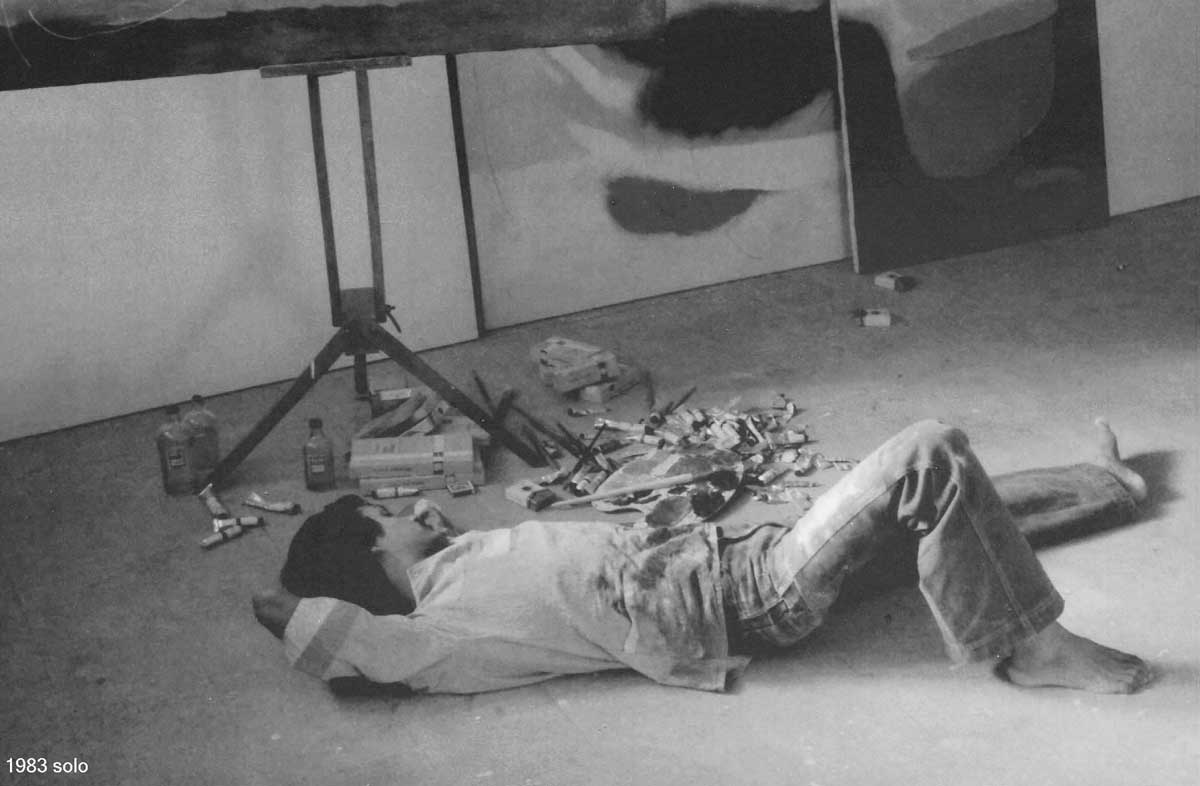 Black and white photograph of Ratnakar Ojha from 1983 showing a person lying on the floor in an art studio, with paint cans, brushes, and canvases visible in the background. The text '1983 solo' appears in the lower-left corner.
